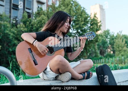 jeune latina jouant de la guitare dans une ville. femme assise sur un banc de pierre pratiquant avec son instrument de musique. Banque D'Images