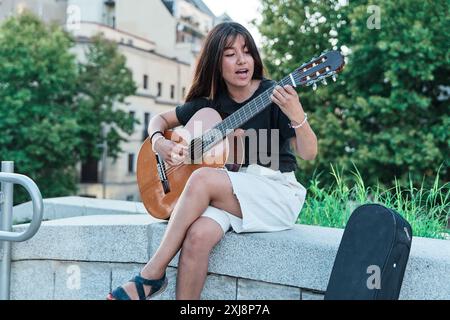 jeune latina jouant de la guitare dans une ville. femme assise sur un banc de pierre pratiquant avec son instrument de musique. Banque D'Images