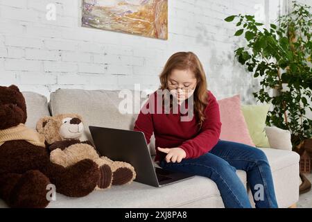 Une petite fille atteinte du syndrome de Down utilise un ordinateur portable sur un canapé, entourée de peluches. Banque D'Images