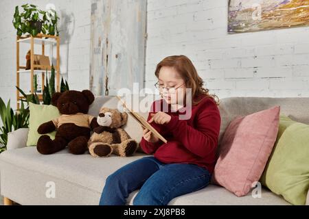 Une petite fille atteinte du syndrome de Down est assise sur un canapé, entourée de jouets en peluche, lisant un livre. Banque D'Images