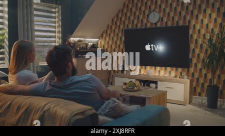 ZAPORIZHYA, UKRAINE - 7 JUIN 2024 : un couple caucasien adulte parle et commence à regarder un film ou une série sur le service de streaming Apple TV assis sur un canapé et se reposant à la maison. Logo Apple TV affiché sur le grand écran du téléviseur. Concept de style de vie. Banque D'Images