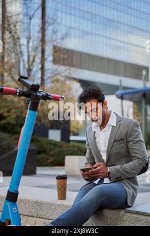 jeune homme d'affaires utilisant son téléphone intelligent assis sur un banc de pierre avec le scooter électrique à côté de lui et des immeubles de bureaux en arrière-plan Banque D'Images