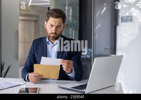 Un homme d'affaires concentré dans un costume lit attentivement un document tout en étant assis à son bureau dans un bureau lumineux et contemporain. La scène transmet professionnalisme et concentration. Banque D'Images