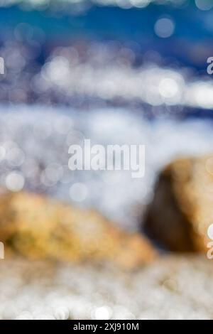 Vagues de mer roulant et s'écrasant sur les rochers avec beaucoup de taches bokeh blanc d'éblouissement du soleil à la surface des ondulations sur l'eau. Vue sur la mer par une journée d'été ensoleillée. Arrière-plan naturel abstrait flou Banque D'Images