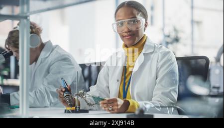 Équipe diversifiée de jeunes scientifiques multiethniques passant un stage dans un laboratoire de haute technologie moderne. Jolie femme africaine regardant la caméra et souriant tout en travaillant avec le fer à souder Banque D'Images