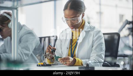 Équipe diversifiée de jeunes scientifiques multiethniques passant un stage dans un laboratoire de haute technologie moderne. Femme africaine travaillant avec le fer à souder portant des lunettes de sécurité et une blouse de laboratoire blanche Banque D'Images