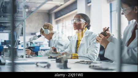 Laboratoire de recherche scientifique moderne à l'université : divers jeunes scientifiques en blouses blanches à l'aide d'un microscope, de cartes de circuit de soudage et de micropuces pour un projet technologique à l'école Banque D'Images