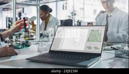 Ordinateur portable avec écran de programme technologique debout sur une table dans un laboratoire de classe scientifique. Divers étudiants pratiquant la soudure de composants électroniques pour leurs projets technologiques Banque D'Images