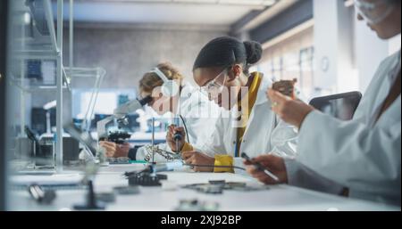 Salle de classe de sciences de l'école secondaire : groupe multiethnique diversifié d'étudiants expérimentant avec des composants informatiques, des cartes de circuit à souder, en utilisant des microscopes. Enseignant marchant et observant l'expérience Banque D'Images