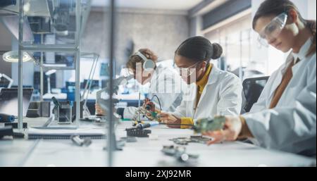 Équipe d'étudiants en sciences multiethniques menant des expériences électroniques dans un atelier universitaire. Divers jeunes camarades de classe utilisant des microscopes, travaillant avec le fer à souder pour créer une carte de circuit imprimé Banque D'Images