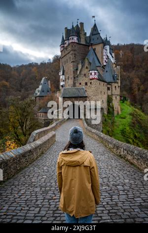 Vue arrière d'une jeune femme avec veste jaune regardant le château historique médiéval d'Eltz dans un paysage d'automne avec des arbres au lever du soleil, Wierschem, Rhinelan Banque D'Images