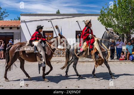 Un jeune gaucho dans un poncho traditionnel rouge Salteño monte avec son frère dans un défilé à Cachi, en Argentine. Les guardamontes en cuir de vache protègent le cavalier Banque D'Images