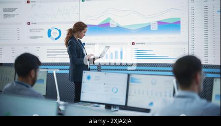 Chef de projet féminin utilisant un ordinateur portable et analysant des graphiques d'information sur grand écran numérique dans le bureau de surveillance. Femme hispanique travaillant pour Big Data Company, divers employés derrière des ordinateurs de bureau Banque D'Images