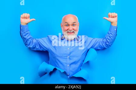 Heureux homme barbu regarde à travers le trou dans le papier bleu. Vieil homme souriant dans une chemise élégante montrant les pouces vers le haut. Casser le papier. Espace de copie pour le texte ou le slogan. Banque D'Images