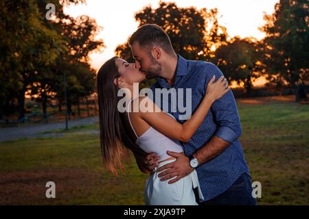 Un homme et une femme s'embrassent et s'embrassent dans un parc au coucher du soleil. Ils sont debout sur un champ herbeux avec des arbres en arrière-plan. Banque D'Images