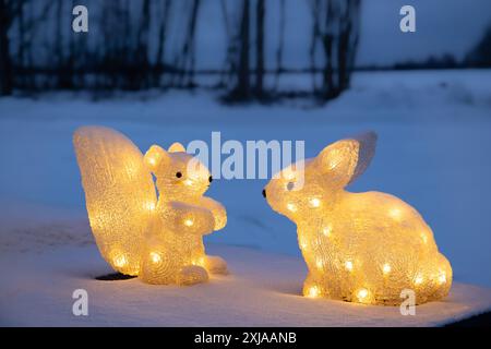 Décorations de Noël illuminées, deux animaux lapin et écureuil jaune lumière la nuit à l'extérieur. Paysage d'hiver enneigé sur fond. Banque D'Images