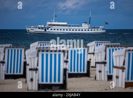 Rostock, Allemagne. 17 juillet 2024. Des chaises de plage vides se dressent par temps changeant sur la plage de Warnemünde sur la côte de la mer Baltique tandis que le bateau d'excursion 'Baltica' part pour un aller-retour. Avec des températures d'une vingtaine de degrés, du vent et de brèves averses de pluie, le temps estival dans le nord de l'Allemagne montre son côté plutôt hostile. Crédit : Jens Büttner/dpa/Alamy Live News Banque D'Images