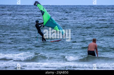 Rostock, Allemagne. 17 juillet 2024. Un surfeur décolle de sa planche pour une balade sur la mer Baltique par temps changeant sur la plage de Warnemünde. Avec des températures d'une vingtaine de degrés, du vent et de brèves averses de pluie, le temps estival dans le nord de l'Allemagne montre son côté plutôt hostile. Crédit : Jens Büttner/dpa/Alamy Live News Banque D'Images