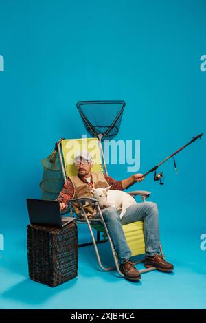 Homme se relaxant sur une chaise longue avec un chien, en utilisant une canne à pêche, un filet de pêche et un ordinateur portable sur une table en osier, placé sur un fond bleu. Banque D'Images