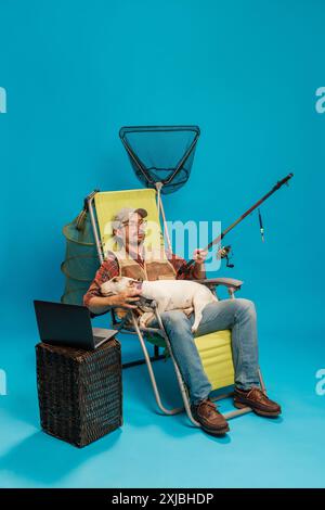 Homme en chemise à carreaux et casquette, se prélassant avec une canne à pêche et un chien, un filet de pêche derrière, et un ordinateur portable sur une table en osier sur fond cyan Banque D'Images