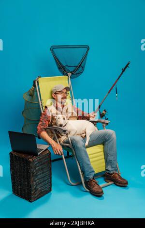 Homme dans une chaise longue, tenant une canne à pêche et un chien, avec un filet de pêche derrière et un ordinateur portable sur une table en osier sur fond cyan Banque D'Images