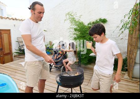 Père et fils grillant ensemble dans un cadre confortable arrière-cour tandis que d'autres membres de la famille se détendent et prennent un verre, créant une guêle familiale chaleureuse et décontractée Banque D'Images