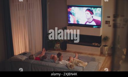 ZAPORIZHYA, UKRAINE - 7 JUIN 2024 : vue en angle élevé de la mère et des enfants allongés sur le canapé avec un chien dans un salon confortable et regardant une émission de télévision ou un film sur Netflix à la télévision. La famille caucasienne se repose à la maison le soir. Home Cinéma. Style de vie. Banque D'Images