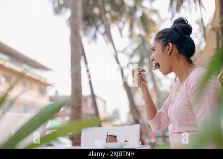 Plaisir du goût. Jeune femme en robe rose à la peau foncée aime le dessert sucré, est assise à table. Banque D'Images