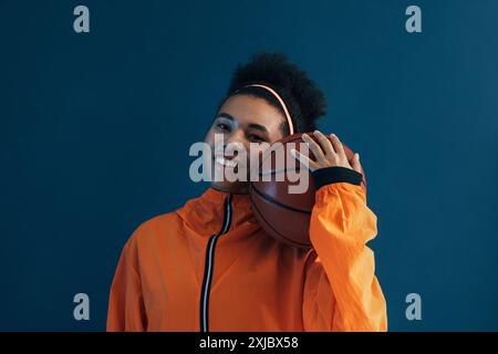 Femelle positive avec cheveux bouclés tenant le basket-ball en studio. Portrait d'une femme souriante posant en studio avec un basket-ball portant des vêtements de sport orange. Banque D'Images