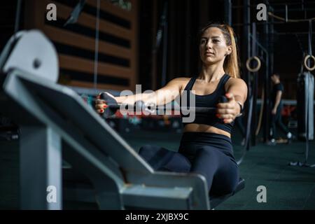 Une femme dans un soutien-gorge de sport noir et des leggings travaille sur une machine à ramer dans une salle de sport. Banque D'Images