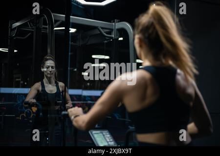 Dans une salle de sport moderne, une femme portant un soutien-gorge de sport noir et des leggings utilise un rameur. Elle est concentrée sur son entraînement et semble déterminée. Banque D'Images
