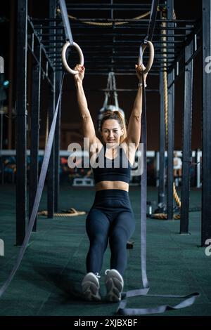 Une femme en vêtements d'entraînement noirs est suspendue à des anneaux de gymnastique, regardant directement la caméra. Banque D'Images
