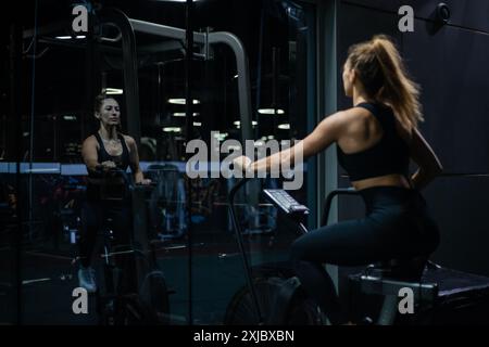 Une femme en vêtements d'entraînement noirs s'entraîne sur un vélo stationnaire dans une salle de sport moderne avec de grandes fenêtres et des miroirs. Banque D'Images