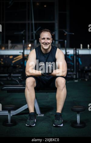 Un homme est assis sur un banc dans un gymnase, portant des vêtements d'entraînement noirs et tenant des poids noirs. Banque D'Images