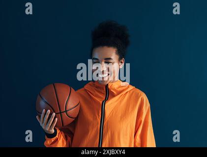 Jeune femme souriante en tenue de fitness orange avec basket-ball. Femme joyeuse avec des cheveux bouclés sur fond bleu tenant le basket-ball. Jeune souriant f Banque D'Images