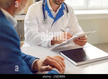 Médecin consultant le patient senior masculin gros plan, écrivant du papier presse-papiers, fixant les symptômes, se plaint Banque D'Images