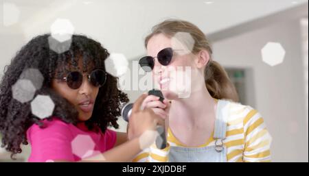 Image de lumières sur des adolescentes heureuses diverses avec des lunettes de soleil et des microphones chantant à la maison Banque D'Images