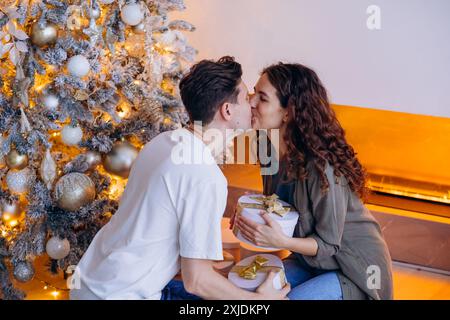 Couple partageant un baiser par un arbre de Noël joliment décoré, tenant des cadeaux emballés et profitant de la saison des fêtes de fin d'année. Homme et femme ont échangé des cadeaux de Noël dans une atmosphère chaleureuse. Banque D'Images