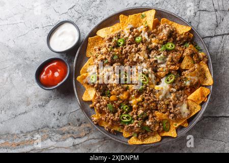 Nachos de maïs avec beaucoup de viande hachée en compote, fromage cheddar et piment jalapeno servis avec des sauces en gros plan sur l'assiette sur la table. Dessus horizontal Banque D'Images