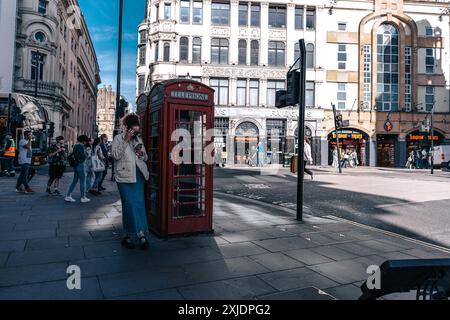 Londres, Royaume-Uni - 10 octobre 2023 : Une femme dans une veste blanche utilise son téléphone tout en se tenant debout à côté d'une cabine téléphonique rouge dans une rue de Londres. Les bâtiments Banque D'Images