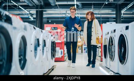 Une femme d'âge moyen interagit avec le conseiller du magasin en électronique domestique lors de la sélection de la machine à laver. Le client senior explore les options d'appareils de blanchisserie. Shopper dans un magasin de détail moderne Banque D'Images