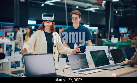 Femme aînée ayant une conversation avec un vendeur dans un magasin d'électronique domestique tout en essayant un appareil de réalité virtuelle. Le client souhaite acheter un cadeau. Le client explore les dernières options avec Expert Banque D'Images