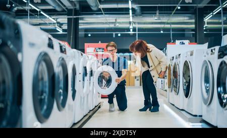 Une femme d'âge moyen interagit avec le conseiller du magasin en électronique domestique lors de la sélection de la machine à laver. Le client senior explore les options d'appareils de blanchisserie. Shopper dans un magasin de détail moderne Banque D'Images