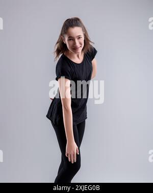 Jeune femme ludique respire le style dans une tenue noire, avec une queue de cheval chic et un sourire confiant dans un décor de studio gris Banque D'Images