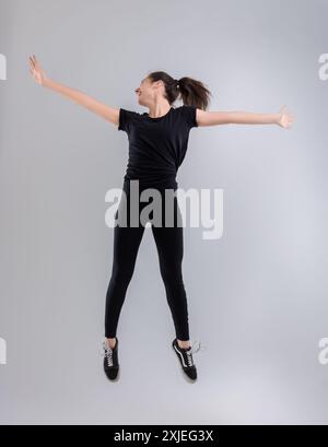 Une jeune femme portant des vêtements d'athlétisme noirs saute joyeusement en l'air avec ses bras écartés, montrant un sentiment de liberté et d'athlétisme Banque D'Images