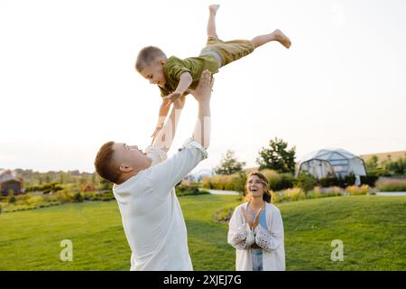 Un père jette son jeune fils en l'air avec un grand sourire au visage. Le garçon rit en volant dans les airs. La mère regarde et applaudit son ha Banque D'Images