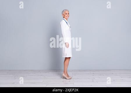 Photo pleine longueur de charmante dame senior heureuse médecin spécialiste qualifié porter une robe blanche isolée sur fond de couleur grise Banque D'Images