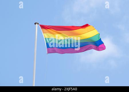 Drapeau arc-en-ciel LGBTQ volant du pôle contre le ciel bleu pour célébrer le mois de la fierté LGBTQIA Banque D'Images