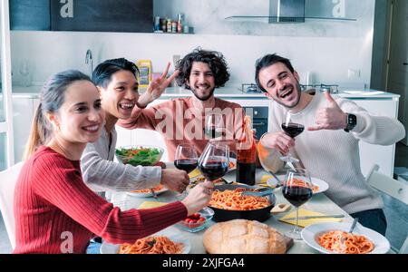 Heureux groupe ethnique d'amis mangeant des pâtes et buvant du vin à la maison déjeuner partie Banque D'Images