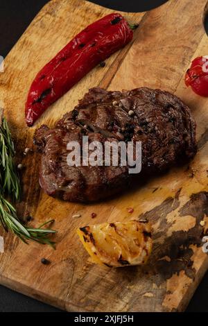 Vue de dessus de Rib Eye Black Angus grillé servi avec du poivre et des herbes sur une planche de bois sur fond sombre. Banque D'Images